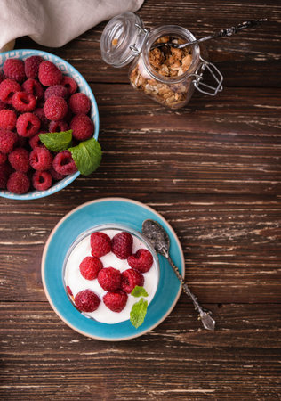 Layered yogurt with granola and raspberries. In a glass, decorated with mint leaves. On dark rustic wooden table background. Healthy food concept.の写真素材