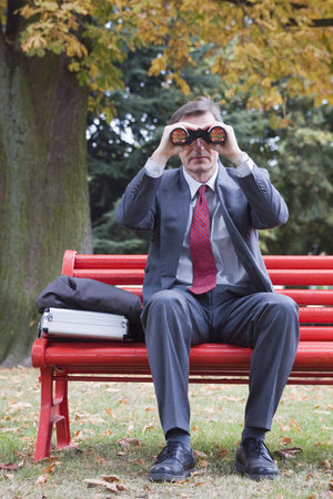 Businessman on a red park bench searching with binocularsの写真素材