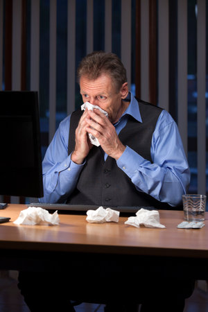 Sneezing mature businessman at a desk in front of a monitor in a dark blue officeの写真素材