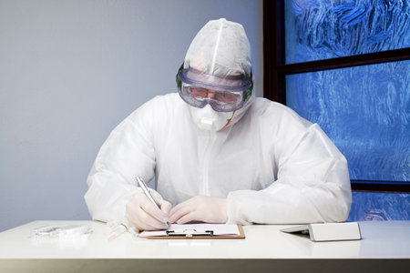 Doctor with protective clothing because of coronavirus writing at a desk in a medical practice