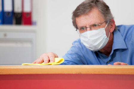 Mature businessman with medical mask cleaning a desk in an office because of coronavirus - selective...