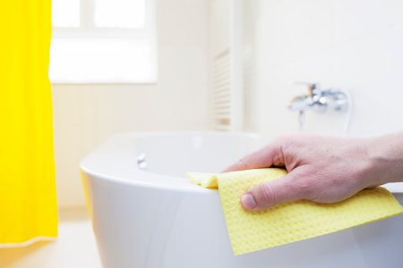 Hand cleaning a tub with a yellow wipe in a bright bathroom - focus on the handの写真素材