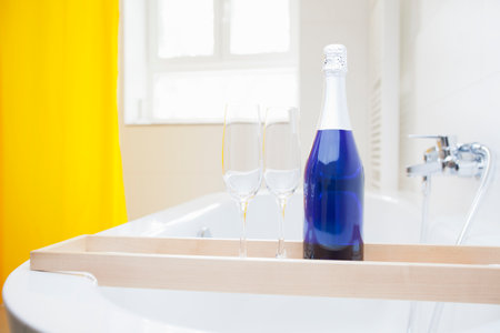 Blue bottle of champagne with two glasses on a bathtub in a bright and clean bathroom - focus on the bottleの写真素材