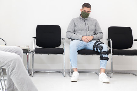 Young man with face mask sitting in a waiting room of a hospital or office - in the foreground the legs of another personの写真素材