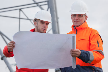 Young and mature architect with hard hat looking at a construction plan at a construction site or oil platform - selective focus on the personsの写真素材