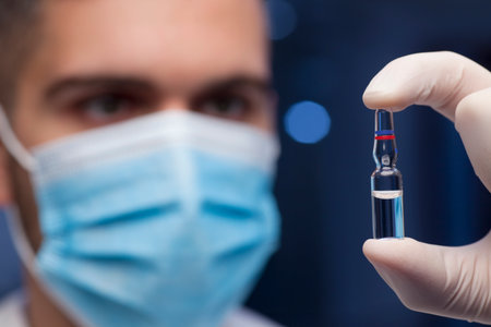Doctor or scientist with medical mask holding an ampule with vaccine in his hand - focus on the ampuleの写真素材