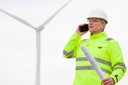 Mature engineer talking on mobile phone in front of a wind turbine with blueprint in his hand - focus on the faceの写真素材
