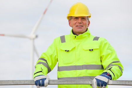Smiling mature engineer with hard hat and protective clothing in front of blurred wind turbine in the backgroundの写真素材
