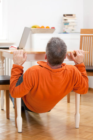 Mature man doing pull-ups at home under a dining table with laptop - focus on the headの写真素材