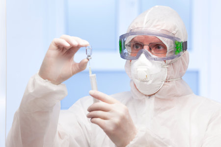Doctor in white protective clothing and medical mask preparing a syringe for vaccination - focus on the faceの写真素材