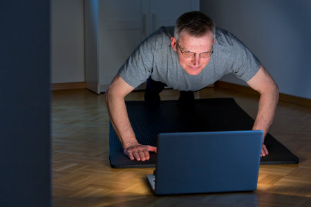 Mature or senior man doing push ups in a dark room at home in front of laptop - focus on the faceの写真素材