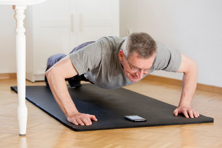 Mature man doing push ups at home with smart phone on the mat - selective focus on the face