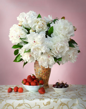 Gorgeous lush white peonies with green leaves in porcelain vase with Chinese painting on openwork tablecloth with golden pattern and dishes with sweet red strawberries and juicy dark burgundy cherriesの写真素材