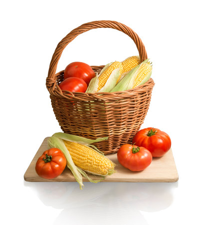 Vegan home kitchen still life of healthy diet products. Ripe golden corn cobs and large red salad tomatoes in old wicker basket on chopping wooden board isolated on white backdrop with clipping maskの写真素材