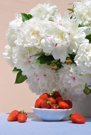 Vivid red juicy sweet strawberries in saucer on the background of a lush bouquet of peonies in delicate white vase with space for text on blue tableclothの写真素材
