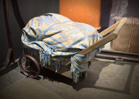 Rough big aged gone hand carry wagon wain lift pile of household items, covered an eldest tattered decorative patterned blanket. View closeup with space for text on night bygone train station backdropの写真素材