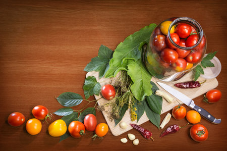 Different varieties of fresh ripe tomatoes arranged in glass jar, knife, leaves, tufts of dry fennel seeds, cloves of garlic, hot pepper isolated on wooden table with space for text. Top view close-upの写真素材