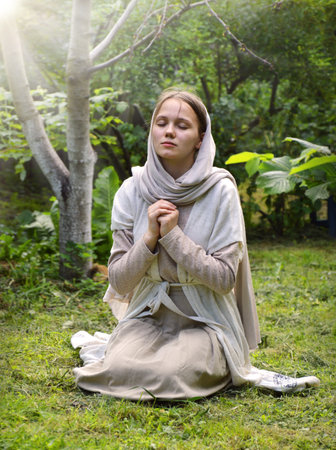 Lone holy young sad east jew Mary old retro shawl veil kneel park lawn hand think ask beg wish Lord Jesus Christ. Rural village teen child kid lady happy joy face thank praise trust peace sun lightの写真素材
