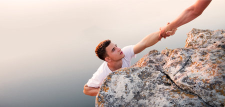 Close up of two risky young alpinist job leader guy showing unity, arm care, trying to rise, trust, hope near a lake, sea, or river. One hangs while the other asks for aid, life, fall, crisis, raise effort, win, reach hill top peak, overcoming abyss, gap, sky, sun.の写真素材