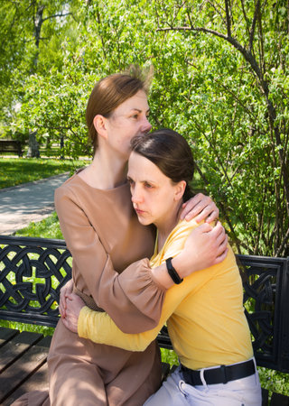 2 young adult lady sit hand talk old urban summer city town garden tree. Teen youth bad feel guilty shame offend think lost love mourn grief pain mood sunny outside nature tell advice pray god faithの写真素材