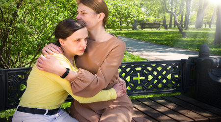 2 young adult lady sit hand talk old urban summer city town garden tree. Teen youth bad feel guilty shame offend think lost love mourn grief pain mood sunny outside nature tell advice pray god faithの写真素材