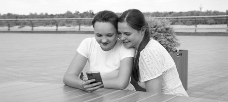 2 young age lady sister sit meet table feel excit surf hand type call app outside summer old city park seat sky text space. teen youth human face eye see sunny happy cheer joy amaz fun emot relax restの写真素材