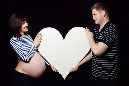 Pregnant Woman and Man Holding a Big White Heart Shape and Looking at each other. Happy Loving Family. Maternity and Paternity Conceptの写真素材