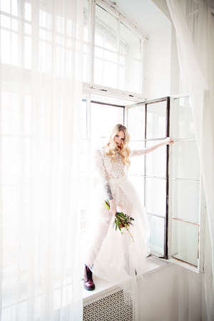 Beautiful Young Blonde Fiancee with Bridal Hairstyle, Event Makeup and Flowers Opening the Window. Fashion Model Woman wearing White Dressの写真素材