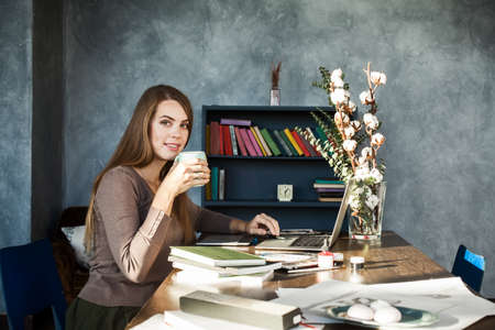 Beautiful Happy Smiling Young Designer Woman sitting at Home Office Desk with Cup of Coffee, posing, looking at camera. Cheerful Model Using Laptopの写真素材