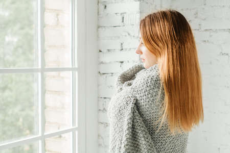 Pretty Redhead Woman Looking out the Window at Home with Grey Woolen Blanketの写真素材