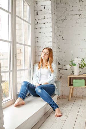Cute girl with red hair sitting in shorts on sunny window in white apartment.の写真素材