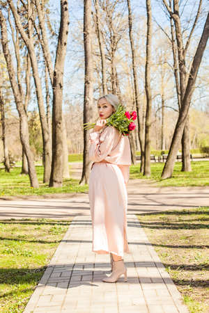 Perfect woman in pink silky raincoat and ankle boots holding bunch of flowers, fashion outdoors portraitの写真素材
