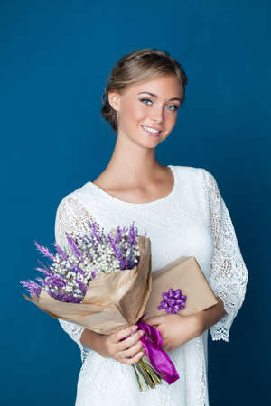 Young friendly woman smiling and holding gift and flowers. Young happy female model in white dress on blue backgroundの写真素材