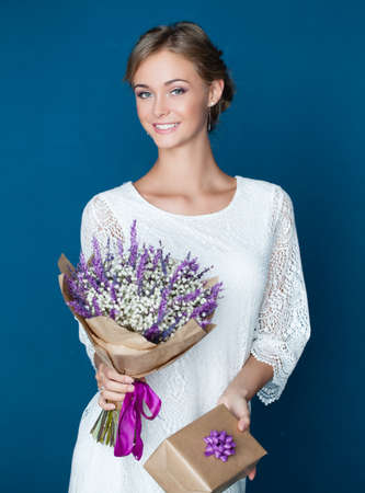 Beautiful smiling woman with gift and flowers. Young happy female model in white dress on blue backgroundの写真素材