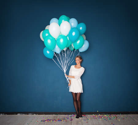 Happy Young Woman in White Dress Holding Balloons on Blue Holiday Background with Confettiの写真素材