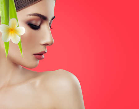 Close up of beautiful young woman's face with healthy skin and tropical flower. Portrait of beautiful model with natural make up. Spa, skincare and wellness concept. Red coral background with copyspace.の写真素材
