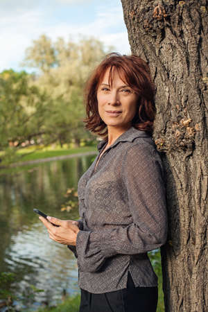 Mature woman with smartphone relaxing in park outdoors. Woman cell phoneの写真素材