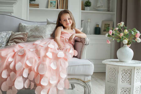 Smiling child girl in pink dress sitting on sofa at homeの写真素材