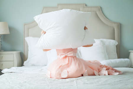 Funny child girl hiding behind a pillow at homeの写真素材