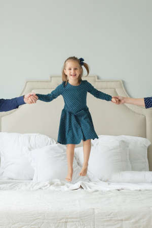 Happy child girl jumping on the bed and holding parents handsの写真素材