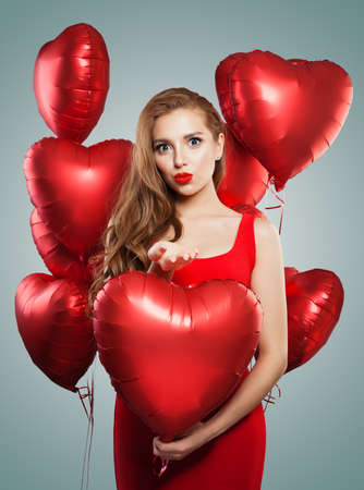 Excited young woman blowing kiss and holding balloons red heart. Beautiful girl with red lips makeup, long curly hair and cute smile. Surprise, valentines people and Valentine's day conceptの写真素材