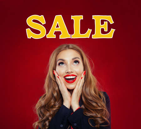 Happy surprised girl looking at sale text on red banner background. Beautiful woman with red lips makeup and long curly hairstyle, sale conceptの写真素材