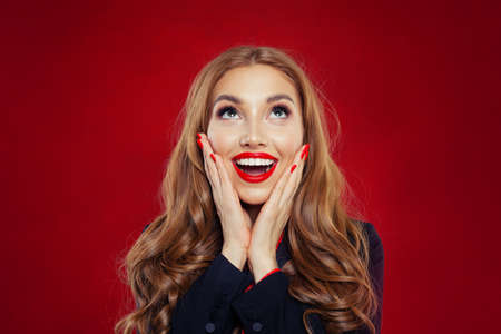 Beautiful surprised woman with red lips makeup and long curly hair. Happy girl looking up on empty red background with copy space. Positive emotions, facial expressionの写真素材