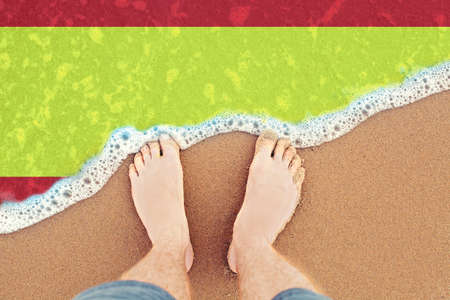 Feet on the sunny sandy beach with flag Spain. Top View on surf.の写真素材