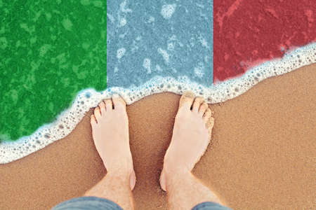 Feet on the sunny sandy beach with flag Italy. Top View on sea surf.の写真素材