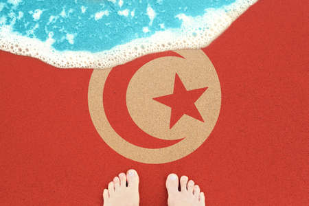 Two feet on the sunny sandy beach with flag Tunisia. View from top on surf.の写真素材