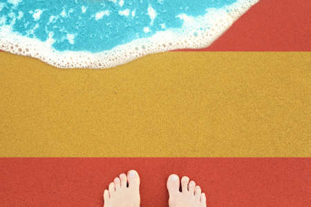 Feet on sea sand and surf. Vacation on ocean beach, summer holiday with flag Spainの写真素材