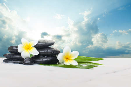 Tropical flowers and black stones ready for healthy therapy and clouds with sunlight. Health objects on white marble tableの写真素材