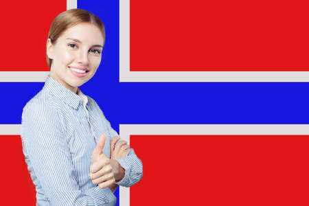 Cheerful brunette woman showing thumb up on the Norway flag background. Live, work, education and internship in Norwayの写真素材