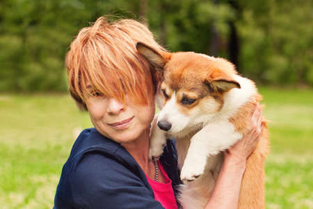 Happy senior woman with her pet friend dog puppy cuddling on green grass in summer park portraitの写真素材